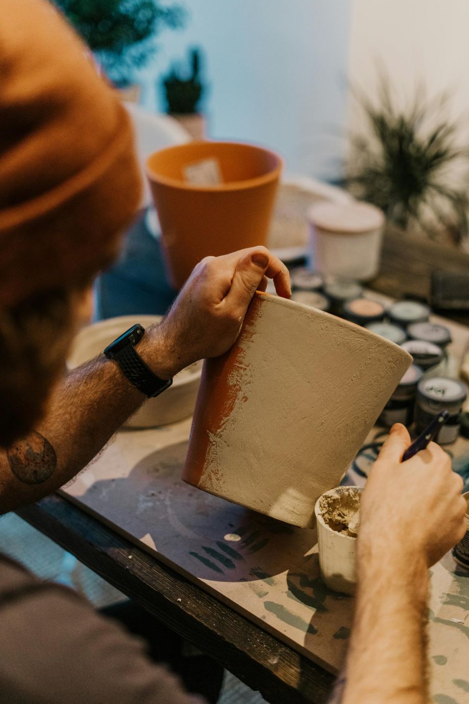 Person painting a flower pot white at a table with art supplies and plants in the background.