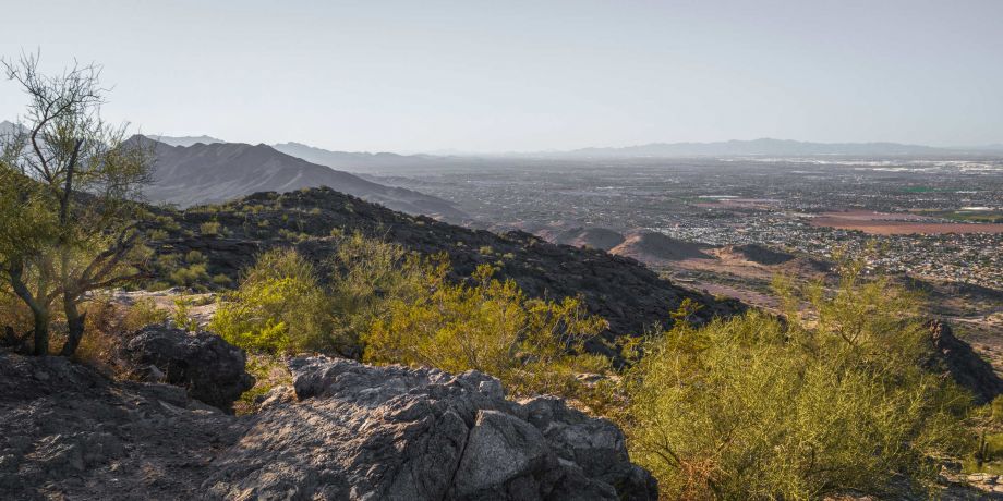 Rocky desert landscape with sparse shrubs, mountains in the distance, and a sprawling city under a clear sky.